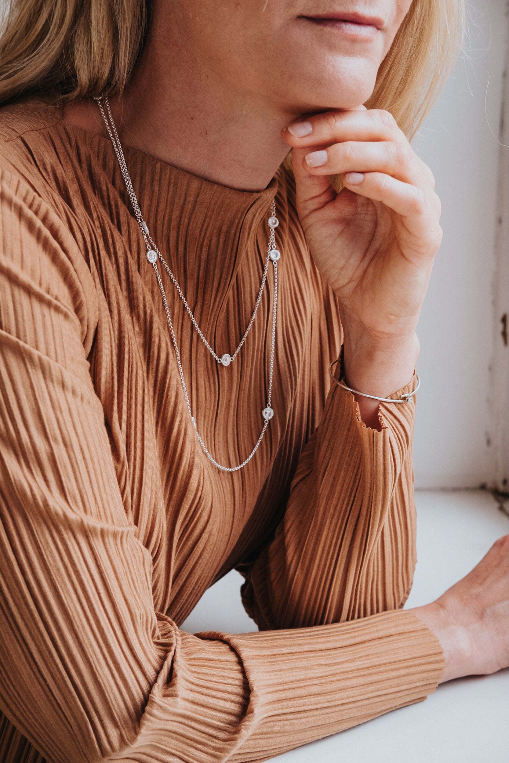 Jag vet inte vem personen är, men bilden visar en kvinna i en plisserad brun tröja som bär ett dubbelt halsband med små hängen.